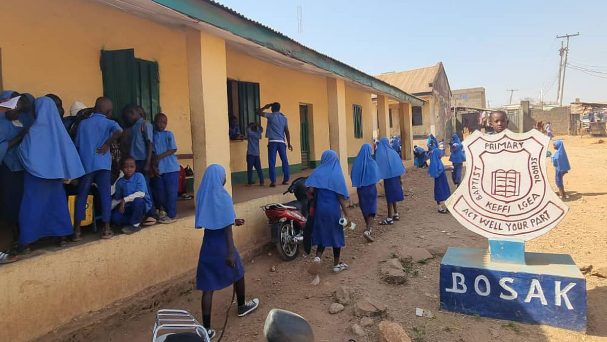 Schoolyard scene with students in blue uniforms, a building, and a school sign.