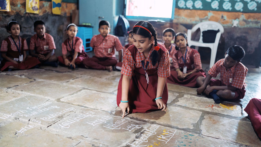 A girl writes on the classroom floor with chalk while surrounded by seated classmates in red plaid uniforms.